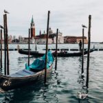 Laguna veneta al tramonto, con riflessi dorati sull'acqua e barche a vela in lontananza.